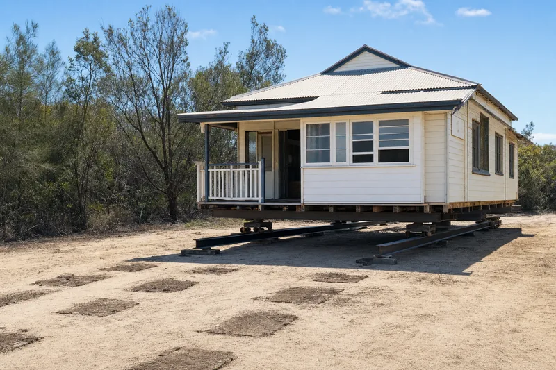 Timber cottage on steel skid beams and rollers part-way through a slide across a flat sandy block, illustrative example