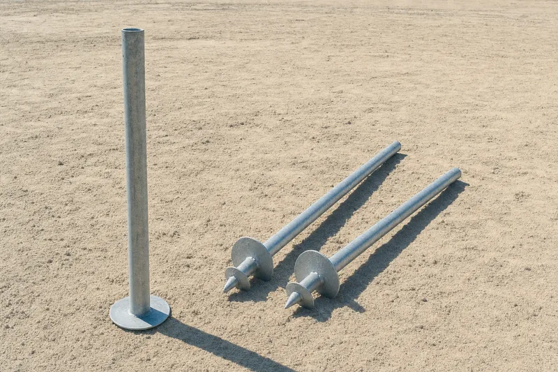 Galvanised screw piers with helical plates, one wound into sandy ground and two laid on bare sand beside it, illustrative example