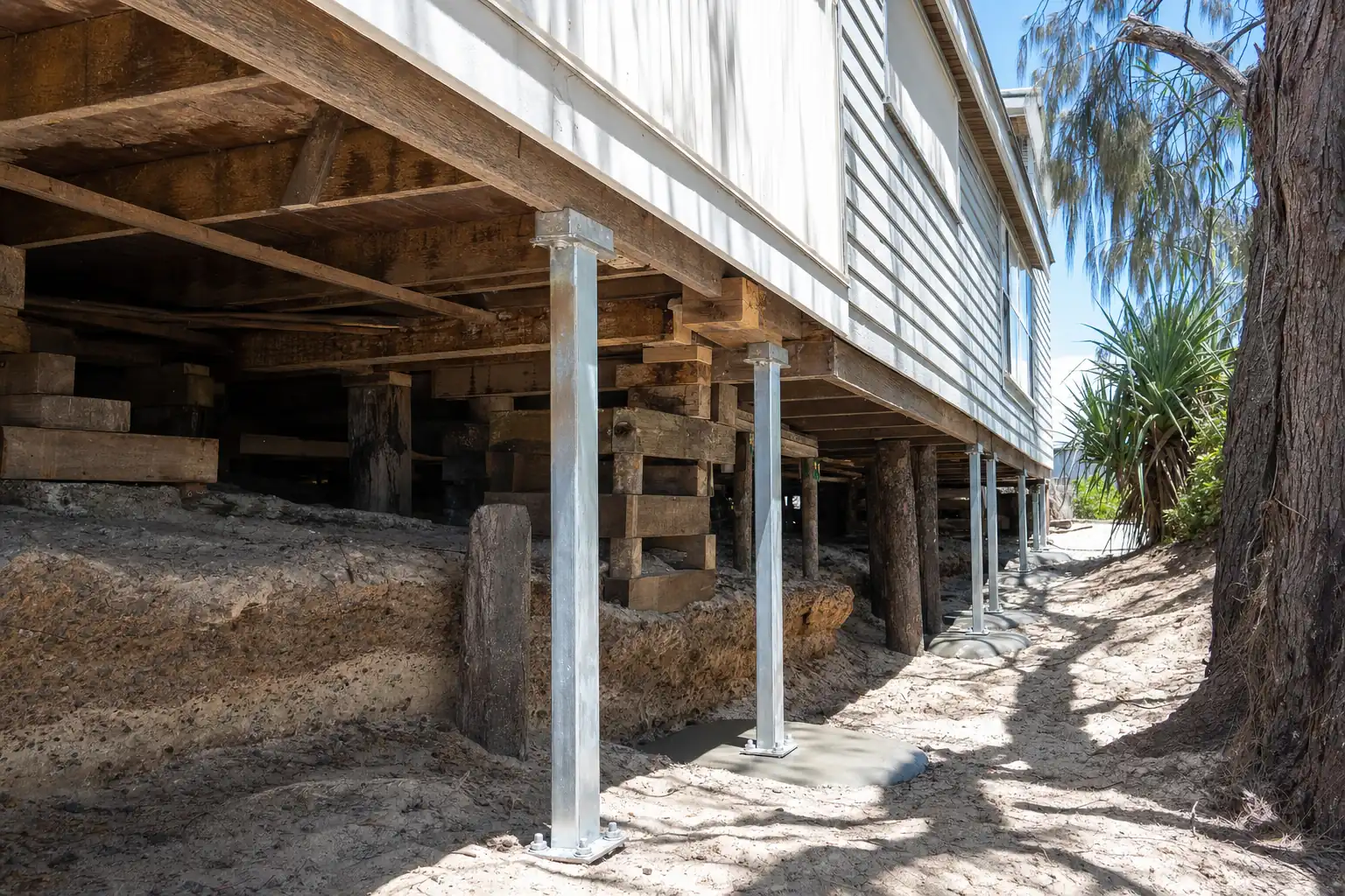 Low-set fibro and weatherboard beach cottage lifted on hardwood cribbing and screw jacks, new galvanised steel stumps standing beside the old timber stumps in coastal sand, illustrative example